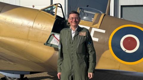 Smiling pilot stands in front of a brown Spitfire. The plane is outside two large grey hangers at Southampton International Airport