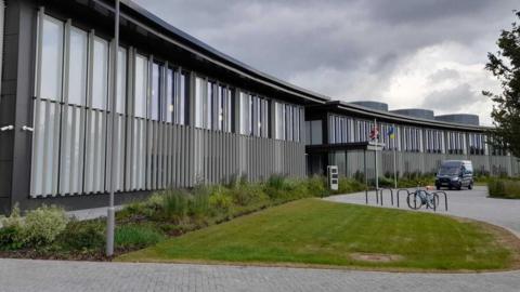 New Shire Hall in Alconbury Weald, Cambridgeshire is a slightly curved crescent shaped building with grey exterior walls and light grey stripes with windows on the front. There are two flags, Great British and Ukrainian, flying at the front. This is the base for Cambridgeshire County Council.