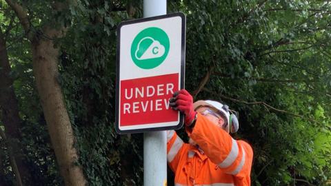 A man wearing an orange jumpsuit and a white helmet up a ladder is removing a road sign that reads "Under review".