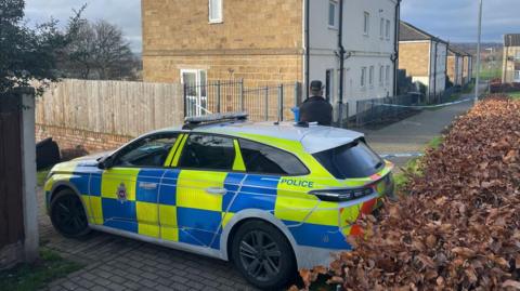 A police car with a police officer stood in front of the vehicle. There is a hedgerow on the right of the image and a police cordon is in the background.
