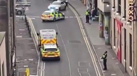 An aerial view of The Cowgate shows several police cars and a police van. There is police tape and two police officers standing guard.