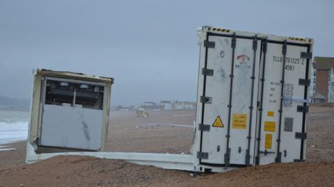 A white container stuck in sand on a beach. The top and sides of the container are missing. The water appears rough and the sky is grey and stormy.