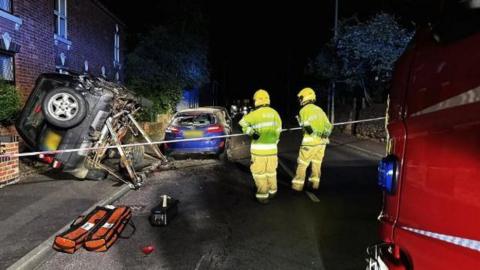 A Land Rover 4x4 vehicle is on its side, propped up against a wall, as two firefighters look on. The image was taken at night, and the blue lights of a fire engine partially illuminate a row of houses to the left of the image.