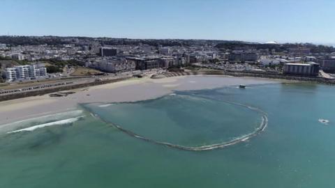 An aerial shot of the Victoria Marine Lake in St Helier. A sandy beach is behind a water-filled the pool, with St Helier behind that. 