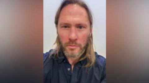 Justin Rundle looks towards the camera. He has long brown hair and a beard. He is wearing a blue shit. 
