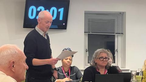 An older bald man wearing a black v-neck jumper with a checked shirt beneath it reads from a piece of paper as he stands up at a council meeting. There is a woman sat beside him with grey hair, wearing a black jacket and black glasses. A woman wearing a blue headwrap can be seen sitting on the bench behind. Behind the man's head is a screen with a timer, showing two minutes and one second of the council meeting has passed.