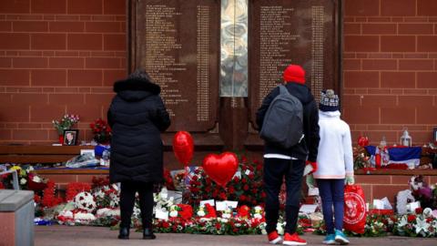People stand in front of the Hillsborough Memorial outside Anfield Stadium in Liverpool. There are flowers and heart balloons in front of a plaque with the names of the 96 victims of the disaster.
