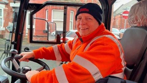 Ricky, a man in his 60s, sits in a clear-sided road sweeping vehicle. He wears an orange high-visibility jacket and a black woolly hat. He is smiling.