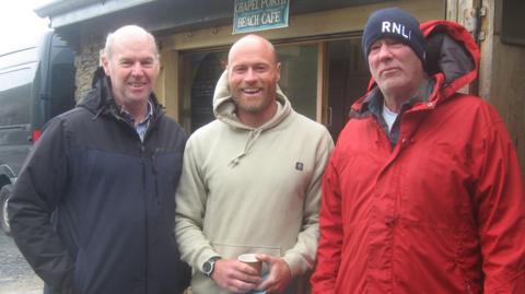 David Lewington (left) RNLI Supervisor Martyn Ward (right) and his son Joshua smile at the camera in coats and hoodies while stood in front of the Chapel Porth Beach Cafe 