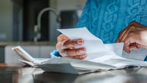 Close up shot of a person looking at bills and receipts on a table next to a calculator in their kitchen. They are wearing a blue jumper and the kitchen sink is in the background.