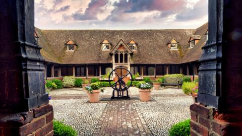 The almshouses are an old building with four sides and a cobbled central area. They have a steeply pitched roof with ornate dormer windows and a covered walkway around the bottom with white doors leading off from it. The central court is nearly planted with benches in the corner.