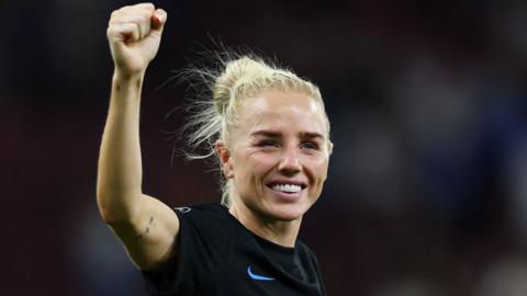Alex Greenwood, with blonde tied back hair and wearing a black England shirt, raises her right hand in the air in a fist and smiles