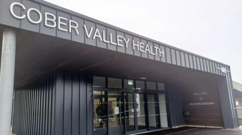 The front of a grey building with automatic doors. It says ' Welcome to Helston Gateway' on it and 'Cober Valley Health'. The sky is grey.