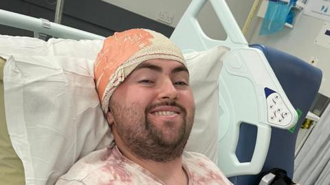 A close-up of a man in a hospital bed. He has head bandages and is smiling.