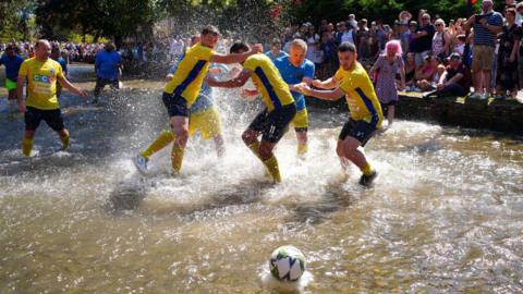 Footballers create splashes in the water of the River Windrush during the annual football match on bank holiday Monday. One team is wearing yellow tops and dark blue shorts while the other is wearing blue tops and yellow shorts. The ball can be seen on top of the water in the foreground and in the background spectators are lining both banks of the river