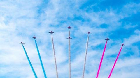 Several planes fly high in formation in the sky. They are spraying blue, white and pink smoke. 