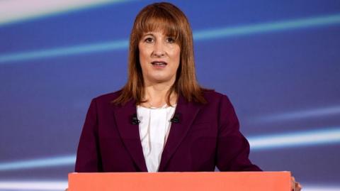 A photohraph of Rachel Reeves standing in front of a blue background. She is wearing a burgundy coloured blazer with a white blouse underneath