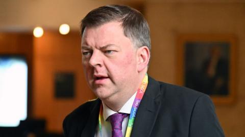 A man with short dark hair, wearing a dark suit, white shirt, purple tie and rainbow-coloured lanyard. He is visible from the shoulders up, with dimly lit walls behind him.