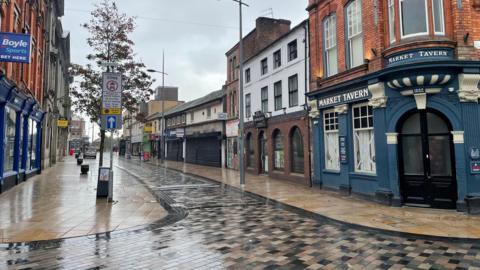 An empty street with wet paving following rain. On the right of the street on the corner is a brick building with blue panelling below reading "Market Tavern". The building has two windows and an arched doorway.