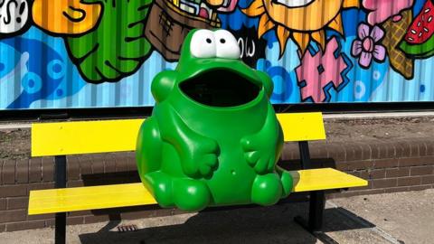 A green bin shaped like a frog placed upon a yellow bench.