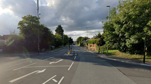 The junction on Painswick Road between Heron Way and Norbury Avenue. There is a pedestrian crossing and a cross roads. There is paths both sides of the carriageway with residential buildings to the left and right.