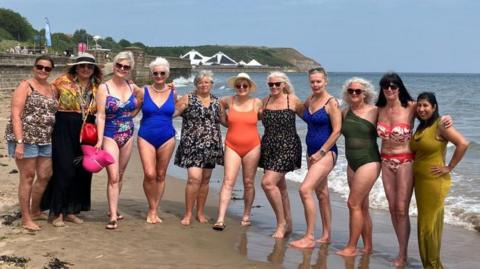 A mixed age group of women pose on Scarborough beach, with the sea, stone sea wall and buildings in the background. Someone women wear swimming costumes and sunglasses, some wear vest tops and shorts, and some wear dresses.
