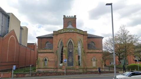 A church building sits next to a large redbrick building on a one-way street