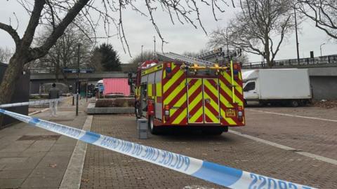 At Milton Keynes Markets a blue and white taped police cordon is in place. The back of a fire engine can be seen within the cordon and so can a white van. The markets sit beneath a bridge and is next to Centre: MK. 