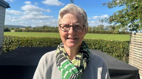 Laura Rowbotham wearing a green and white striped shirt and tortoise shell glasses. She is stood in front of a hedge which looks over a grassy field.
