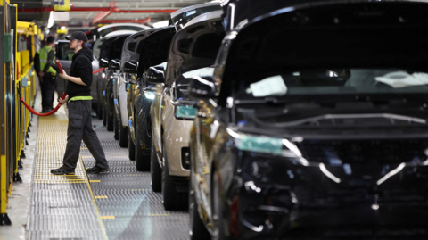 A row of Land Rovers sitting in a factor, to the left of the row of cars is a male worker in dark cargo trousers, a black hooded top and a baseball cap, holding a red cable which looks like it could be involved in the car production process