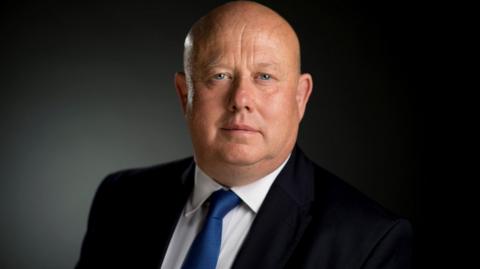 Former council leader Ian Hudspeth.  He is a bald man with blue eyes wearing a black suit, white shirt and blue tie - he is looking at the camera.