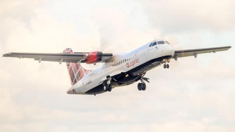 A propeller passenger plane in flight with its wheels down. The aircrfat has Loganair livery, including a red tartan tail.