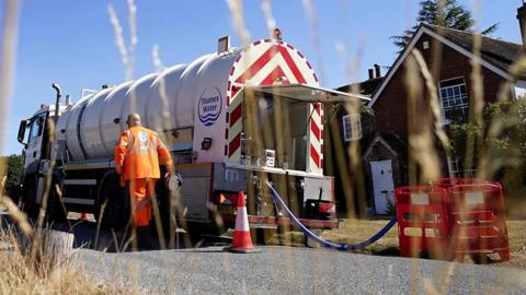 A Thames Water branded water truck parked in front of a house with a blue pipe from the truck leading to some kind  of works being done on a section of the road. A man in hi-viz is returning to the truck.