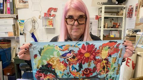 Claire Baker, who has pale pink hair and large, square tortoiseshell glasses, holds up a piece of embroidery showing colourful flowers and birds.