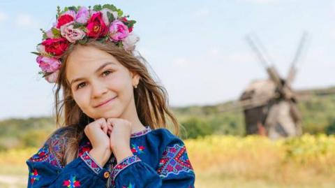 A girl wears Ukrainian national dress and a Vinok wreath of flowers outdoors. Her dress is blue with brightly-coloured patterns and the wreath is pink, red and purple.