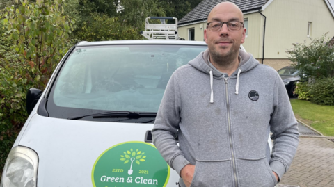 Ian Bowyer wearing glasses and a grey zip-up hoodie and standing in front of a white van with a Green and Clean logo on the bonnet. 