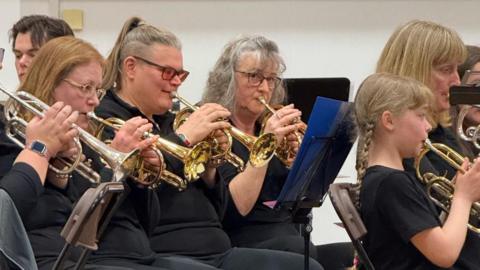 Four women and a girl are all dressed in black playing brass instruments in front of black music stands.