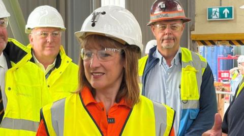 Chancellor Rachel Reeves is wearing an orange top and yellow high vis jacket and a white hard hat. Several men who are wearing similar protective clothing are standing behind her.