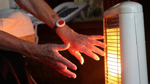 An elderly woman warms her hands at an electric fire