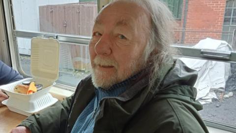 A man with long greying hair past his chin and with a light blonde, white beard looks sideways into the camera, slightly over his left shoulder. He sits by a table which has an open polystyrene box on it with a sandwich and a napkin inside. Behind him is a window with a metal bar across the middle.  