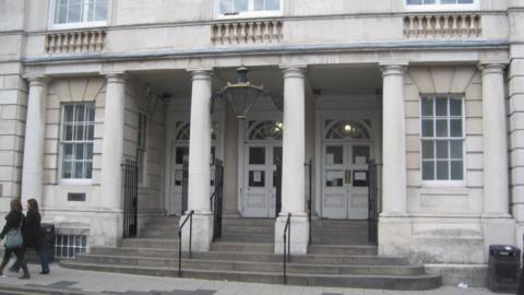 The exterior of Lewes Crown Court. A light grey, stone building with three large white doors. Two people walk right to left on the pavement in front of the building