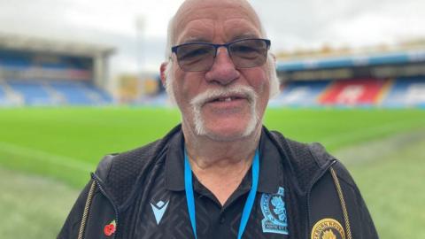 Head and shoulder of Ian Snape standing on the pitch at Ewood Park. He has long white sideburns and a handlebar moustache and wire framed glasses. He is wearing a dark Rovers top and jacket