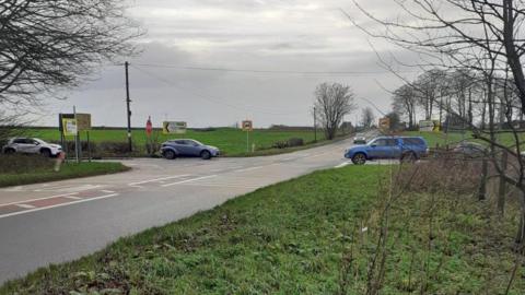 A few cars getting ready to drive through a cross roads. There are fields all around the roads. It is an overcast and grey day.