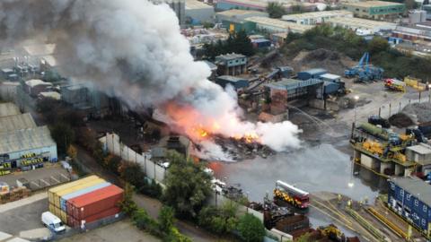 A large plume of thick grey smoke billowing up into the sky from the recycling centre. Flames are coming up from rubble on the ground and water is being sprayed onto the fire. There are several fire engines at the scene. 