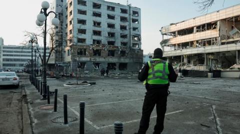 A police officer wearing a yellow vest stands with their back to the camera, looking at a damaged apartment building from across an empty carpark