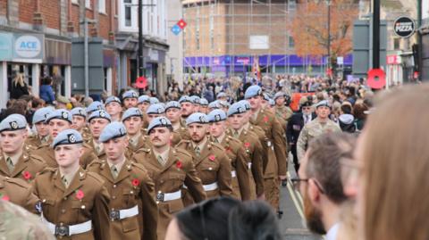 Soldiers wearing poppies marching through a street in Andover on Remembrance Sunday. People are watching them from the side. It is overcast.