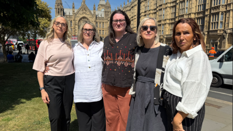 Five women stood holding each other by the waist and facing the camera. They are stood outside Parliament.