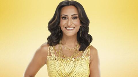 This image shows Strictly Come Dancing contestant Dr Punam Krishan wearing a yellow sequined dress and lots of gold jewellery. She has brown hair and gold eye makeup and is smiling at the camera. The photo has a solid yellow/gold background.