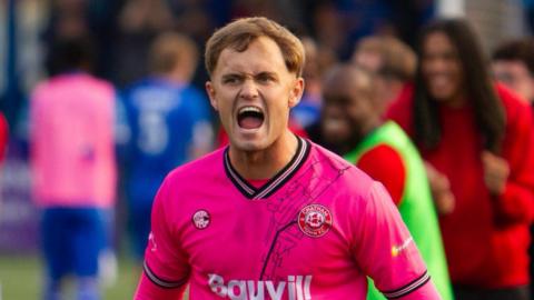 A young man with blonde hair wearing a hot pink football shirt while shouting. Behind him, blurred football players move in all directions.