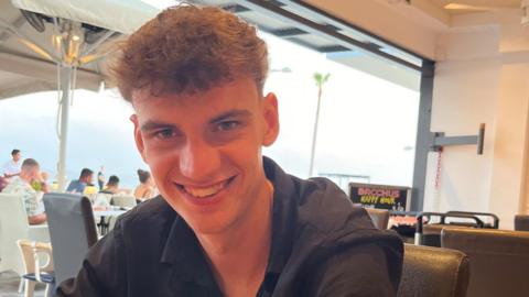 Zain Shaw, a young man with curly brown hair and a black shirt on, smiling at the camera, while sat in a restaurant.
